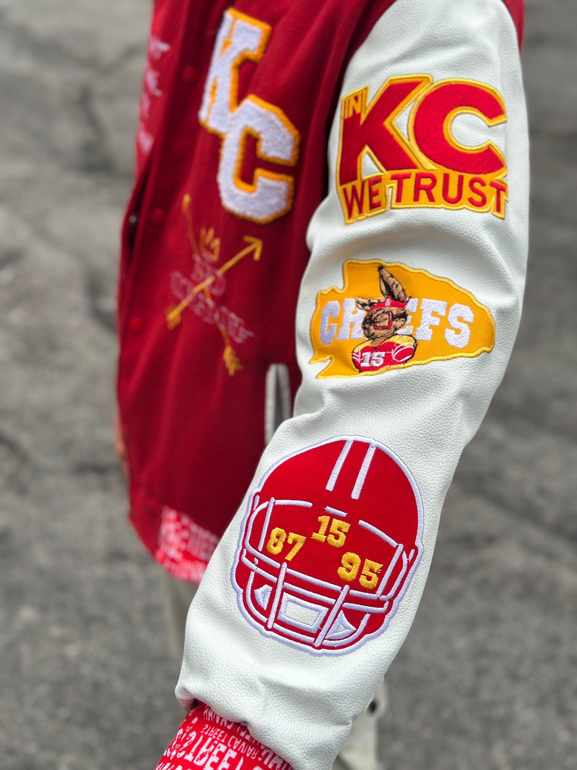 Close-up of Street Caviar KC Chiefs varsity jacket sleeve featuring embroidered helmet patch with numbers 15, 87, 95, “In KC We Trust” patch, and Chiefs player graphic on white faux leather sleeve
