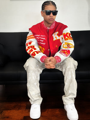 Model wearing Street Caviar red KC Chiefs varsity jacket with embroidered patches, white paisley cargo pants, and white sneakers, seated on a black couch.