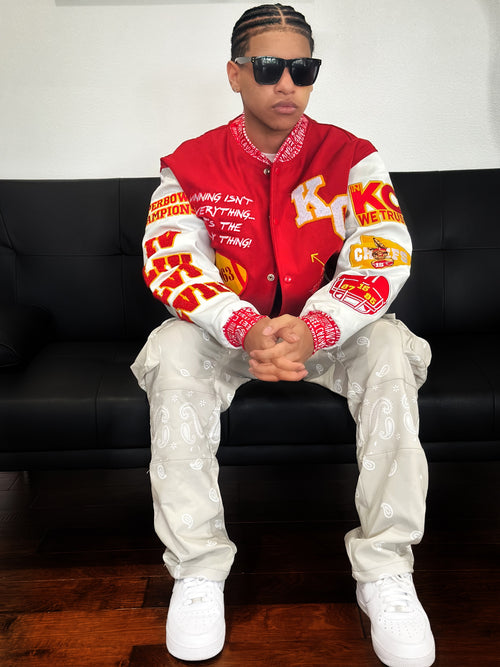 Model wearing Street Caviar red KC Chiefs varsity jacket with embroidered patches, white paisley cargo pants, and white sneakers, seated on a black couch.
